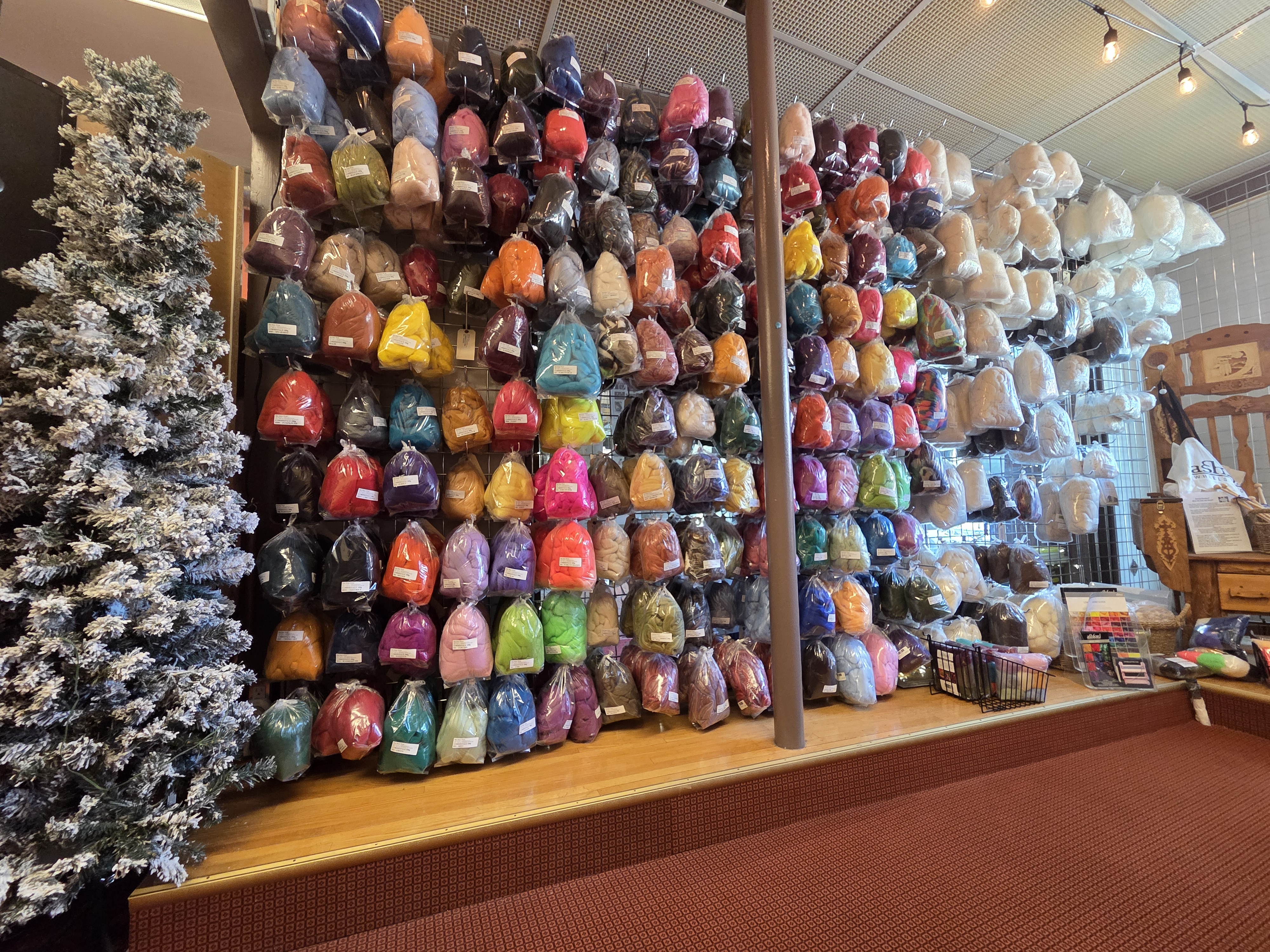 A vibrant display of various colored wool packaged in bags, arranged on a wall in a cozy yarn store, with a frosted Christmas tree visible on the left and a wooden chair on the right.